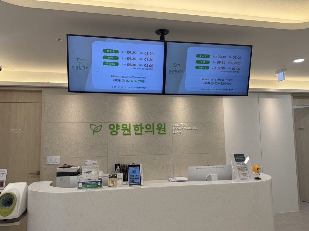 한의원 대기실 상단에 설치된 삼성 디지털 사이니지 2대에 진료시간과 상담전화 안내가 표시된 모습 — 원장님 부재 시에도 환자에게 필요한 정보를 자동으로 전달하는 원내 커뮤니케이션 화면