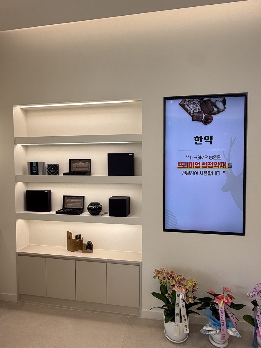 한의원 진열장 옆 벽면에 설치된 세로형 디지털 사이니지 — h-GMP 승인 청정약재 선별 사용을 안내하는 한약 콘텐츠가 표출되고 있는 화면