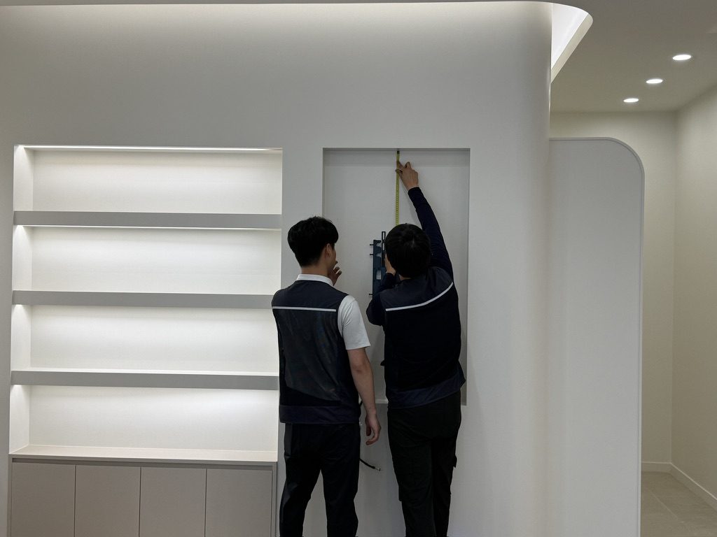 한의원 벽면 매립 사이니지 시공 현장 — 시공 기사 두 명이 벽체에 마련된 매립 공간의 치수를 줄자로 실측하고 브라켓 위치를 확인하는 모습