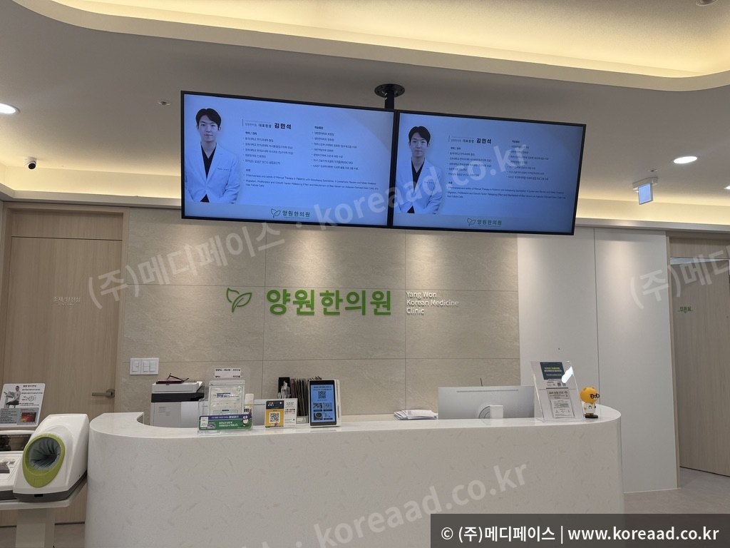 한의원 접수대 위 천장에 사이니지 두 대가 나란히 설치되어 접수 대기 환자에게 원내 안내 콘텐츠를 보여주는 실제 설치 사진
